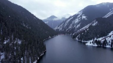 Kolsai Dağı Gölü, kış ormanında. Bulutların, kozalaklı ağaçların, aynadan yansıyan suyun, kardaki tepelerin ve dağların drone görüntüsü. Sarı günbatımı. Tekneler bazı yerlerde yüzer. Kazakistan, Almaty