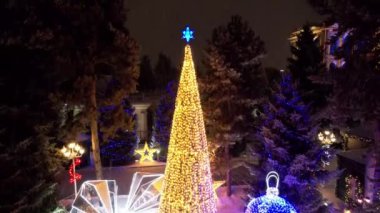 Noel ağacıyla süslenmiş parlak Noel süslemeleri. Noel ağacında çelenkler ve neon oyuncaklar parlıyor. Noel ve Yeni Yıl için dekorasyon. Kar yağıyor. İnsanlar fotoğraf çeker. İHA 'dan üst görünüm