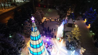 Noel ağacıyla süslenmiş parlak Noel süslemeleri. Noel ağacında çelenkler ve neon oyuncaklar parlıyor. Noel ve Yeni Yıl için dekorasyon. Kar yağıyor. İnsanlar fotoğraf çeker. İHA 'dan üst görünüm