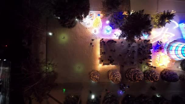 雪の下でクリスマスの装飾は明るく輝いている クリスマスツリー ランタン モミの木 ガーランド さまざまなライトのドローンからのトップビュー 車を渡している 人々は歩いている — ストック動画
