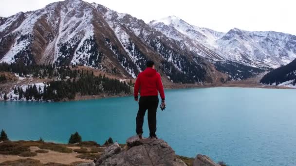 男は高い石の上に山の湖を見下ろす立っている 水の色はターコイズブルーで 太陽の光が表面に輝きます 遠くに雪の山や森のキャップ — ストック動画