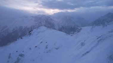 Karlı dağlarda bulutların olduğu mistik şafak. Beyaz tepeler bulutlar ve karla kaplıdır. Dik kayalıklar. Zirvenin zirveleri. Şafak vakti güneş ışınları bulutların arasından geçer.