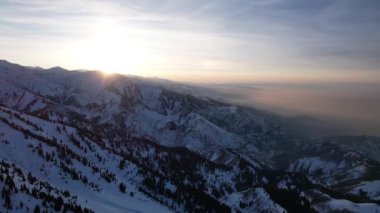 Yüksek karlı dağlarda destansı kırmızı-turuncu gün batımı. Uzun kozalaklı ağaçlar var, tepelerde kar var. Yüksek zirveler. Güneş ışınları bulutların üzerine düşer ve sis vadiden aşağı iner. Almaty