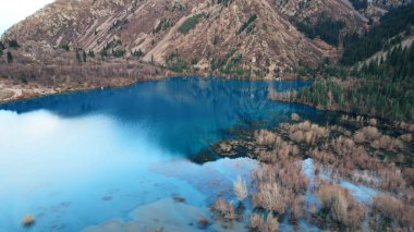 Issyk Dağı Gölü ve Baraj 'ın en üst manzarası. Küçük bir nehir göle akıyor. Tepelerde sarı-yeşil ağaçlar var. Su gökyüzünü yansıtır. İnsanlar kıyı boyunca ve patika boyunca yürür. Kazakistan