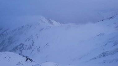 Dağların tepesinde bulutların arasında bir kurtarma kulübesi. Evin yanında duran bir dağcı var. Beyaz bulutlar karlı dağları kapladı. Sarı şafak ve bulutlar. Karlı tepeler. 01.05.2023 Almaty