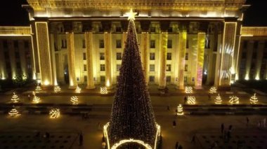 A large Christmas tree in the lights on the square. Top view from the drone on the street with cars and people, a bright building in garlands, various decorations for the new year. Almaty, December