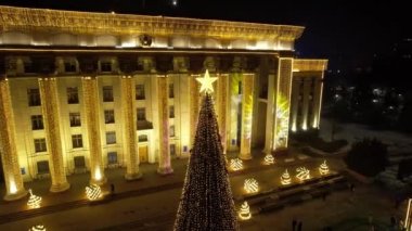 A large Christmas tree in the lights on the square. Top view from the drone on the street with cars and people, a bright building in garlands, various decorations for the new year. Almaty, December