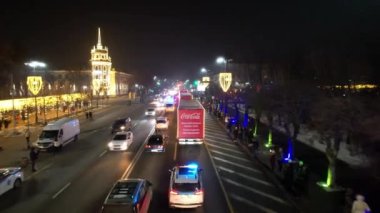 A New Years caravan of Coca Cola cars is traveling along the road. People meet and wave. There is a tall Christmas tree in garlands, lights and decorations are shining all around. Almaty, 12.28.2022