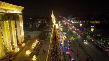 A large Christmas tree in the lights on the square. Top view from the drone on the street with cars and people, a bright building in garlands, various decorations for the new year. Almaty, December