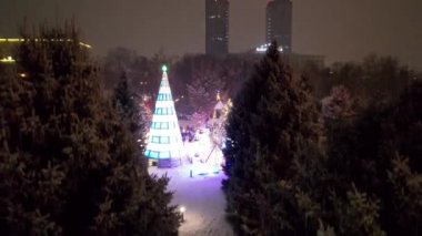 Noel ağacıyla süslenmiş parlak Noel süslemeleri. Noel ağacında çelenkler ve neon oyuncaklar parlıyor. Noel ve Yeni Yıl için dekorasyon. Kar yağıyor. İnsanlar fotoğraf çeker. İHA 'dan üst görünüm