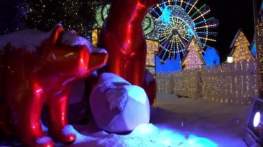 Bright Christmas and New Year decorations. Bears are sitting in white snow, snowflakes are falling. The fence is all lit up. Garlands are glowing all around. Gifts, Christmas trees and a viewing wheel