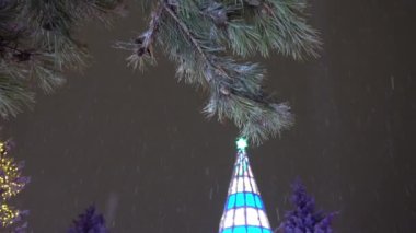 Snow is falling. The Christmas tree is glowing. Coniferous green branches are covered with snow, cones hang down. Christmas decoration - all in garlands. Festive mood. People admire and take pictures