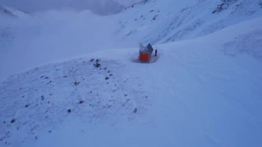 Dağların tepesinde bulutların arasında bir kurtarma kulübesi. Evin yanında duran bir dağcı var. Beyaz bulutlar karlı dağları kapladı. Sarı şafak ve bulutlar. Karlı tepeler, kayalar uzanıyor. Almaty