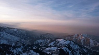 Yüksek karlı dağlarda destansı kırmızı-turuncu gün batımı. Uzun kozalaklı ağaçlar var, tepelerde kar var. Yüksek zirveler. Güneş ışınları bulutların üzerine düşer ve sis vadiden aşağı iner. Almaty