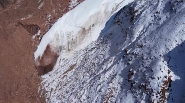 Dağlarda eski bir kar buzulu. Yukarıdan, karla kaplı dik uçurumlardaki bir dronun görüntüsü. Bir buzulla kaplı yüksek zirveler. Bazı yerlerde büyük taşlar var. İzler görünüyor.