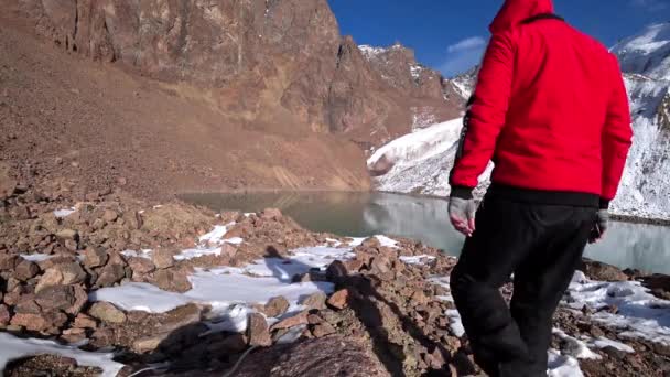 一个穿运动服的家伙去了山上的一个莫兰湖里 在高高的岩石中形成的古老的雪冰川 那家伙戴着手套 穿着红色夹克和黑色裤子 蓝天白云 阿拉木图山区 — 图库视频影像