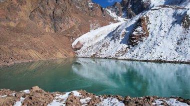 Zümrüt suyla kaplı bir dağ gölü bir buzulu ayna gibi yansıtır. Dağların zirvelerini görebiliyorsun. Göl kısmen donmuş. Bazı yerlerde büyük taşlar ve kar var. Moraine Gölü