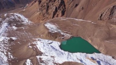 A mountain lake with emerald water is like a mirror. Top view from a drone. The moraine lake is partially frozen. There are large stones and snow in places, a glacier. Pipes and a house are visible