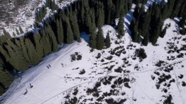 İki dağcı ormanda karlı bir sırt boyunca yürüyorlar. Uzun kozalaklı yeşil ağaçlar. Ağaçların gölgeleri karın üstüne düşüyor. Büyük sırt çantaları taşıyorlar. Bir İHA 'dan üst görünüm