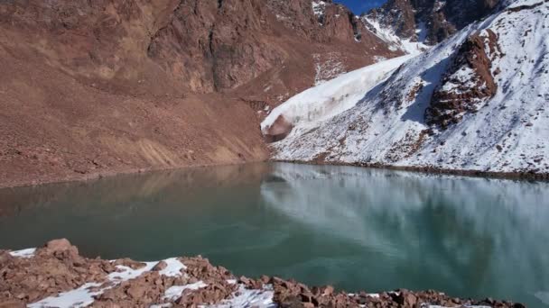エメラルド水のある山の湖は鏡のような氷河を反映しています 山の峰が見えます 湖は部分的に凍結されている 場所には大きな石や雪があります モレーン湖 — ストック動画