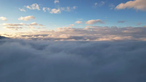 Flying among the ocean of clouds in the mountains. The fog is like big ocean waves crashing against high mountains covered with snow. Spruce trees grow in places. Sunset. Orange rays of the sun