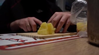 A guy with a knife in his hands is slicing potatoes. Black legs, wooden cutting board. There is a pot. Hiking conditions. A survival hut. A gold wedding ring on her finger. Camping overnight.