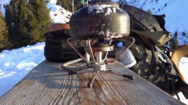 The burner is burning with a blue fire with a kettle. The burner and the cylinder are on the bench. In the background there are green fir trees, mountains in white snow and a beautiful sunset