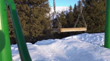 Swing in the mountains with a view of the forest. Metal chains and a wooden board. Good fastening. White snow is lying, coniferous trees are growing, and snow peaks are visible in the distance