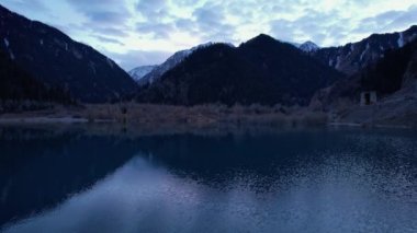 Gün batımında aynalı su ile Issyk Dağı Gölü. Suyun rengi gözlerimizin önünde değişir. Temiz suda ağaçlar var. Karlı dağlar ve yeşil tepeler görülebilir. Bulutlar yansıtılır