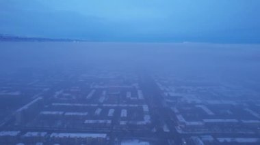 Dawn over the city in fog and smog. Mountain view. A light haze hangs over the city, cars drive, signs glow. Clouds, sun rays and snowy mountains are visible in the distance. Almaty, Kazakhstan