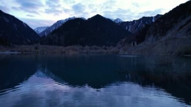 Gün batımında aynalı su ile Issyk Dağı Gölü. Suyun rengi gözlerimizin önünde değişir. Temiz suda ağaçlar var. Karlı dağlar ve yeşil tepeler görülebilir. Bulutlar yansıtılır