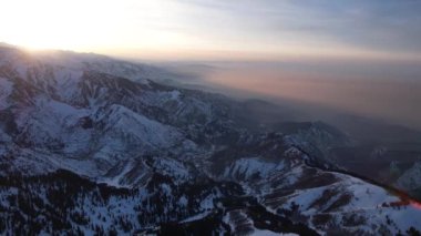 Yüksek karlı dağlarda destansı kırmızı-turuncu gün batımı. Uzun kozalaklı ağaçlar var, tepelerde kar var. Yüksek zirveler. Güneş ışınları bulutların üzerine düşer ve sis vadiden aşağı iner. Almaty