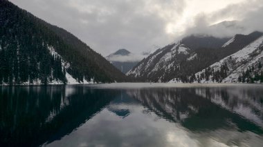 Kolsai Dağı Gölü, kış ormanında. Bulutların, kozalaklı ağaçların, aynadan yansıyan suyun, kardaki tepelerin ve dağların drone görüntüsü. Sarı günbatımı. Tekneler bazı yerlerde yüzer. Kazakistan, Almaty