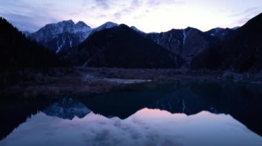 Gün batımında aynalı su ile Issyk Dağı Gölü. Suyun rengi gözlerimizin önünde değişir. Temiz suda ağaçlar var. Karlı dağlar ve yeşil tepeler görülebilir. Bulutlar yansıtılır
