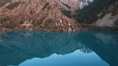 Dağ gölündeki suyun koyu mavi ayna rengi. Düz yüzey bir ayna gibidir, ağaçlar, sarı-yeşil tepeler, dağlar ve gökyüzü yansıtılır. Ağaç gövdeleri suda duruyor. Issyk