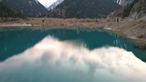 Dark blue mirror color of water in a mountain lake. The smooth surface is like a mirror, trees, yellow-green hills, mountains and the sky are reflected. Tree trunks are standing in the water. Issyk