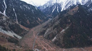 Dağ nehri ve sonbahar ormanı manzarası. Arabalar için toprak bir yol görünür. Ağaçlar sarı-kahverengi-yeşildir. Yüksek karlı tepeler. Nehir geçitten geçiyor. Büyük taşlar var.