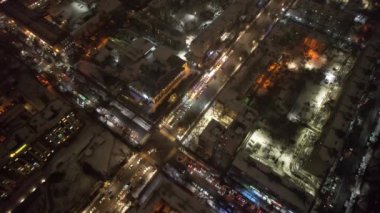 Red big trucks with Coca Cola drive through the streets of the city. Average traffic of cars. Lights and headlights are shining. New Years mood. Top view from the drone on the houses and the road