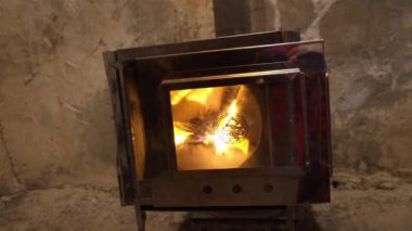 A fire is burning in a camping stove with a glass door. The process of smoldering fuel briquettes is visible. The lining of the stove made of mirror metal reflects the gray old walls of the hut.