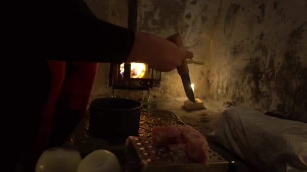 他们在不寻常的野营条件下做饭 一座古老的茅屋 篝火在营房的火炉里燃烧 蜡烛在附近 那家伙把切碎的肉扔进平底锅里 桌子上有个弓 — 图库视频影像
