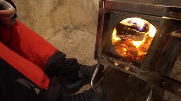 男がキャンプ用ストーブの近くで靴を乾かしている 火は燃えていて 靴から蒸気が出ています 鏡面コーティングを施した金属オーブン 赤いズボングレーの山のブーツ 濡れたトレッキングシューズ — ストック動画