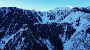 Coniferous forest in snowy mountains. White clouds float across the sky. Top view from a drone. In places, the road to the mountains and buildings are visible. There is a pipe along the gorge. Sunset