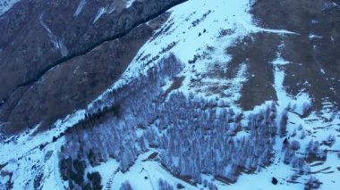 Coniferous forest in snowy mountains. White clouds float across the sky. Top view from a drone. In places, the road to the mountains and buildings are visible. There is a pipe along the gorge. Sunset