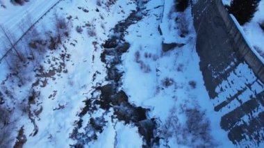 A mountain river runs along a snowy riverbed in the forest. There are big stones. View from above. Animal tracks are visible in places. There is a construction along the river - a concrete wall