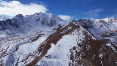 High snowy peaks and a small glaciological station. Drone view of the blue sky with clouds, steep cliffs and rocks. An ancient glacier covered with snow. Small houses are standing. Moraine lake