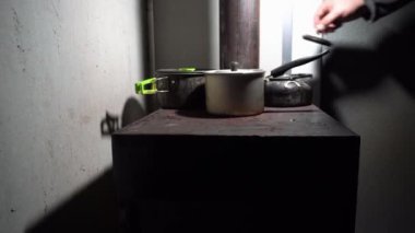 There are pots on a metal stove with a fire. Food is being prepared and snow is being heated. The fire is burning inside. A metal pipe comes out of the roof. A house for survival in the mountains