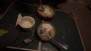 Stir the sour cream with a spoon in a plate with broth and dumplings. Camping food on a small table in an abandoned hut. There is a lid, an estimate and plates with food.