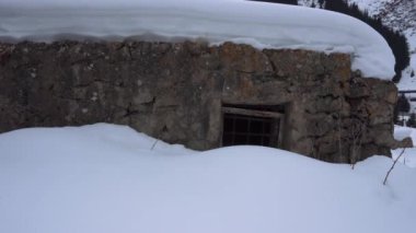 An old stone prison or hut in the mountains. Rusty bars on the windows. The walls consist of huge stones. Dry bushes under white snowdrifts. The roof is covered in snow.