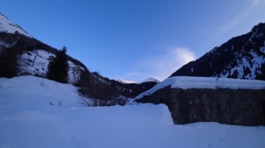 Timelapse of clouds in the mountains and huts. Snowy mountains, forest and clouds against a blue sky. Animal tracks are visible in the snow. An old abandoned hut made of stones. Dry bushes.