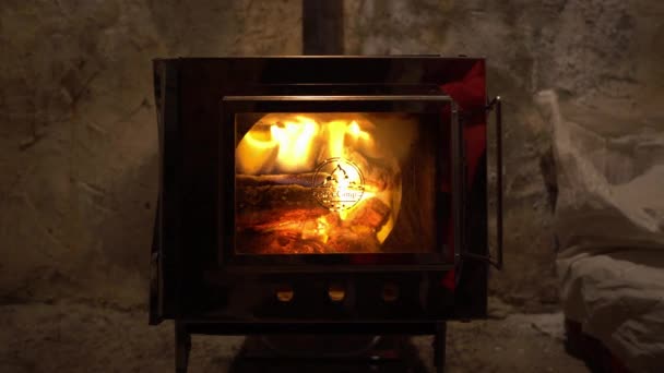 火はガラスの扉でキャンプストーブの中で燃えています 燃料の練炭を製錬する工程が見えます 鏡の金属製のストーブの裏地は小屋の灰色の古い壁を反映しています — ストック動画
